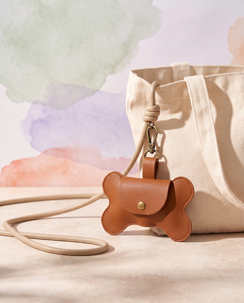 Bone-shaped dog waste bag holder charm clipped to a neutral leash beside a cream tote