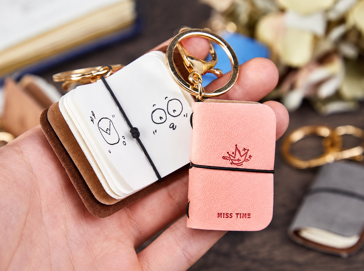 Mini notebooks shown open and closed to illustrate pages, elastic closure and gold-tone attachment; one notebook in the selected color